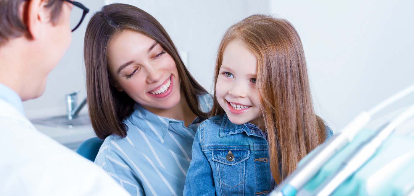 Young girl talking to her dentist while sitting in her mothers lap